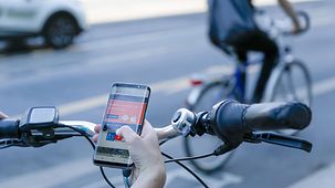 Eine Radfahrerin beugt sich über den Lenker und hält ein Smartphone in der Hand. Aufgerufen ist die Internetseite des Radwegs Deutsche Einheit. Zu sehen ist der Hinweis, dass es eine App zum Radweg gibt. Im Hintergrund ist ein Fahrradweg zu sehen.