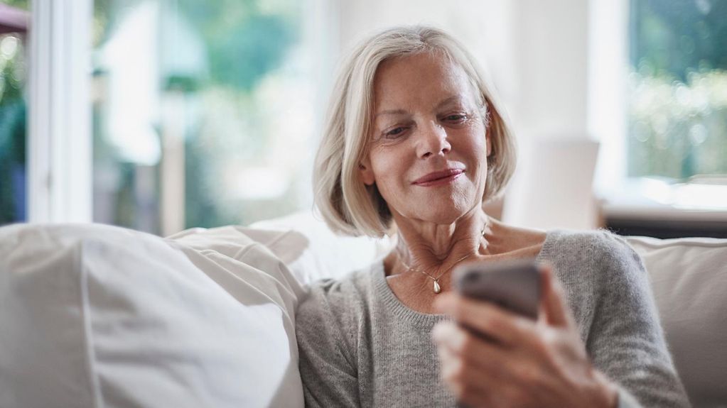 Ältere Frau sitzt in ihrem Wohnzimmer und schaut auf ihr Smartphone.