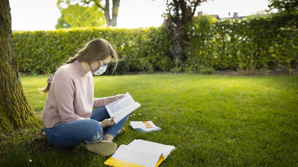 Junge Studentin mit Maske sitzt an einem Baum im Freien und lernt.