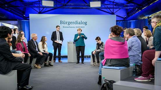 Kanzlerin Merkel steht vor dem Publikum beim Bürgerdialog. Hinter ihr steht vor einer blau beleuchteten Wand eine Leinwand mit der Aufschrift "Bürgerdialog 18. März 2019".
