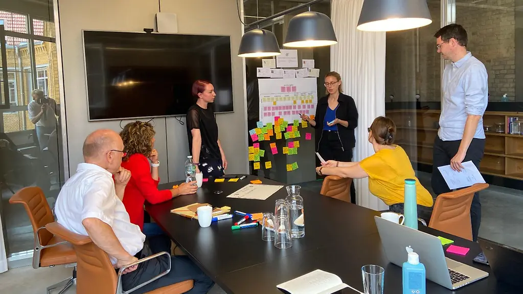 Tech4Germany: Mitarbeiter sitzen an einem Tisch oder stehen vor einem weißen Flipchart und diskutieren.