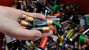 Das Foto zeigt eine Hand mit AA-Batterien über einem Recycling-Behälter für Altbatterien.