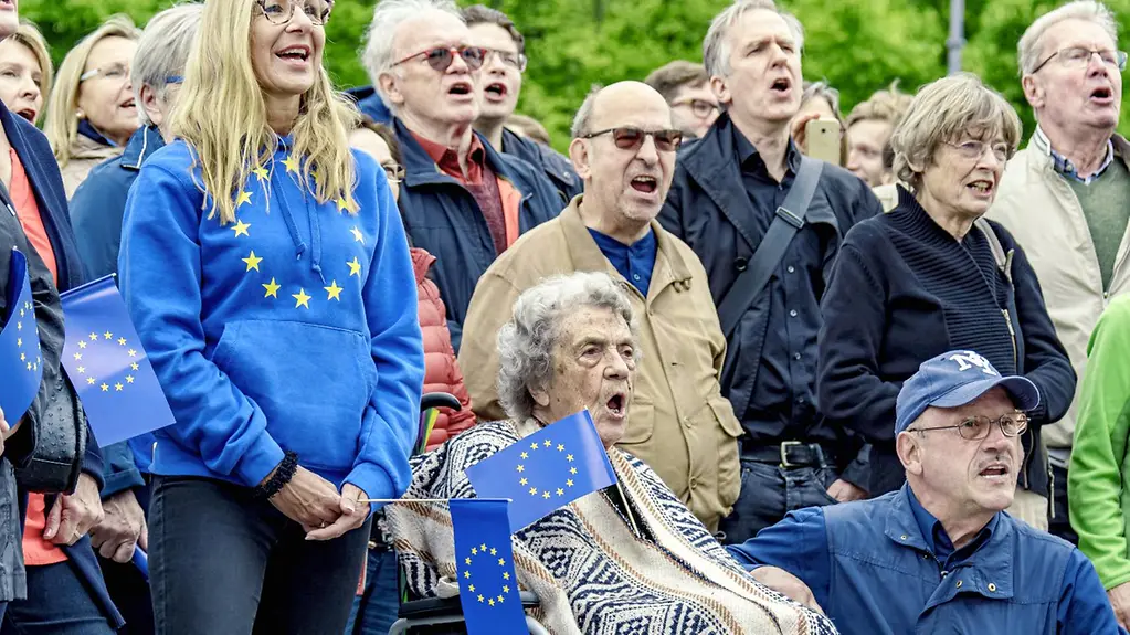 People of all ages with European flags and logos sing together.