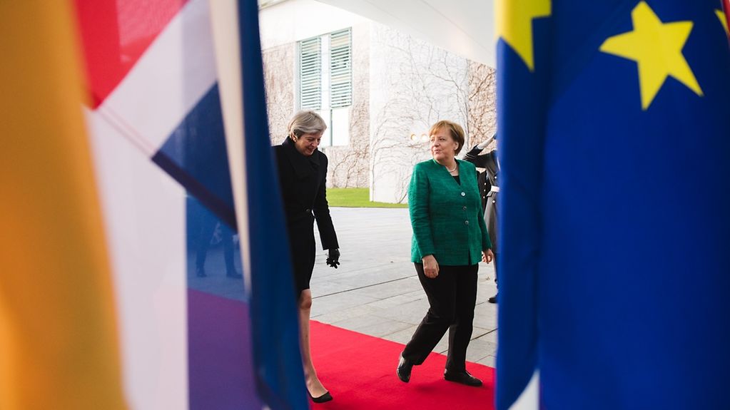 Kanzlerin Merkel und die britische Premierministerin Theresa May gehen umrahmt von der deutschen, der britischen und der europäischen Flagge