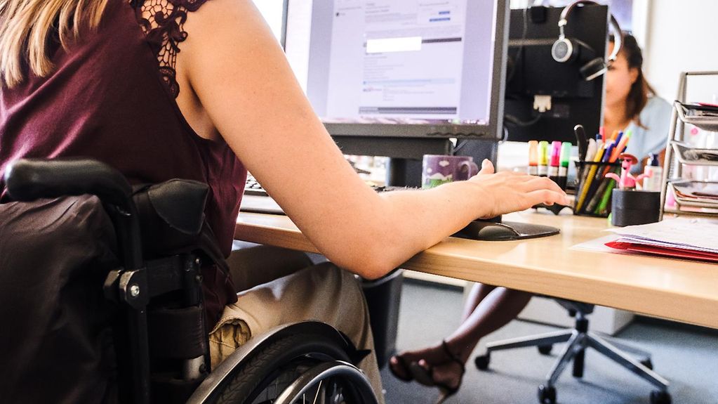 Eine Frau im Rollstuhl sitzt an ihrem Schreibtisch und arbeitet an einem Computer.