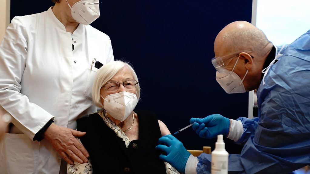 L’image montre une dame âgée se faisant vacciner par son médecin. Un autre médecin se tient à ses côtés.