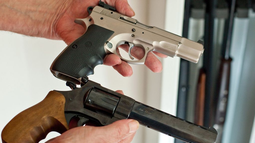 Ein Sportschütze zeigt eine Pistole und einen Revolver vor einem Waffenschrank mit drei Gewehren.