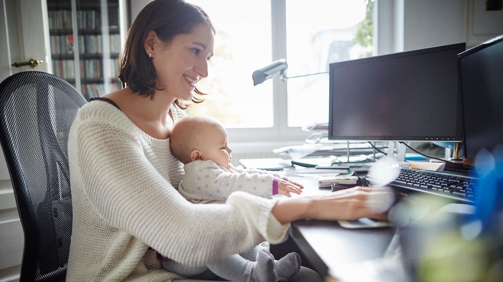 Lächelnde Mutter hält Baby am Schreibtisch im Heimbüro. 