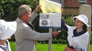 Ein Mann und eine Frau enthüllen ein Schild, auf dem das Wappen der Waldenser und ein historischer Text dazu abgebildet ist.