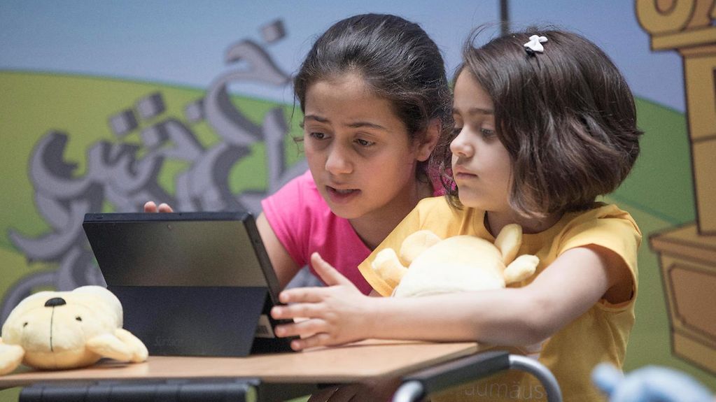 Zwei Mädchen sitzen an einem Tisch und schauen auf einen Tablet-Computer.