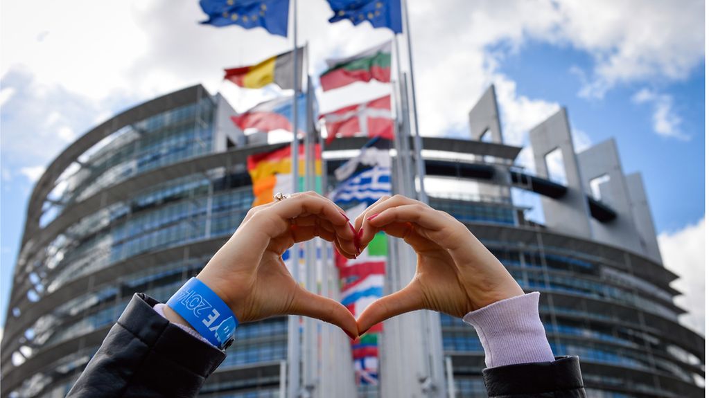 Une femme forme un cœur avec ses mains devant le Parlement européen avec des drapeaux