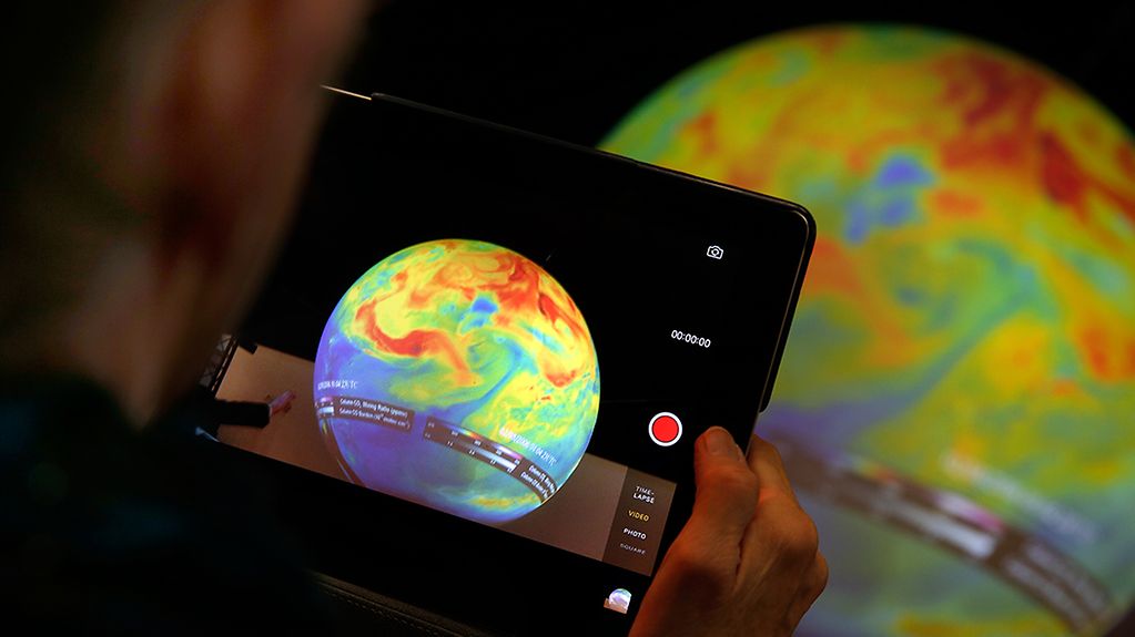 Eine Frau macht mit ihrem Tablet ein Foto eines Globus auf dem farblich die unterschiedliche Belastung durch CO2 Emissionen dargestellt ist.