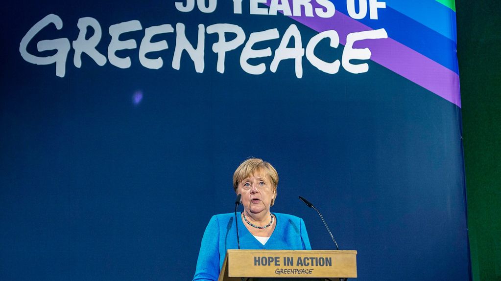 Das Bild zeigt Angela Merkel an einem Rednerpult vor einer blauen Wand. Auf dem Pult steht Hope in Action, Greenpeace