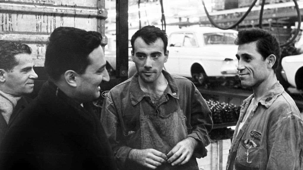 The picture shows several men engrossed in conversation. In the background you can see a conveyor belt with cars that are nearly finished.