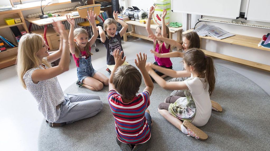 Lehrer und Schüler sitzen in einem Kreis auf dem Boden ihres Klassenzimmers und heben die Hände. 