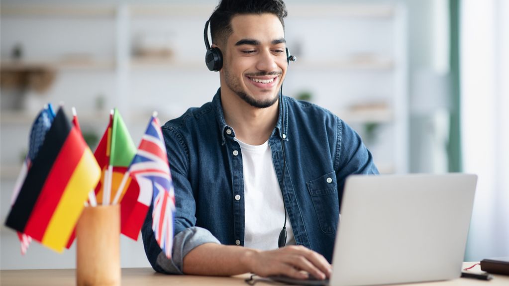 Junger Mann, der online Fremdsprachen lernt, am Schreibtisch sitzt und Laptop, Headset verwendet, auf der Tastatur tippt und lächelt.