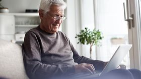 Porträt eines älteren Mannes, der zu Hause auf der Couch sitzt und ein Laptop-Modell benutzt. 