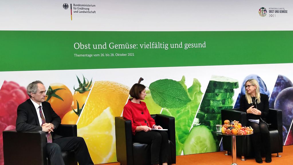 Auf dem Podium sitzen die Teilnehmenden Dr. Thomas Schmidt, Dr. Marianne Altmann und Prof. Monika Schreiner. Im Hintergrund ist Obst und Gemüse abgebildet und der Schriftzug "Obst und Gemüse: vielseitig und gesund" zu lesen.