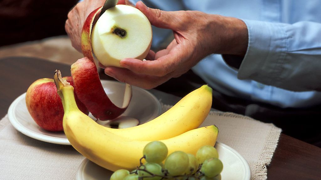 Das symbolhafte Foto zeigt die Hände eines älteren Menschen beim Schälen eines Apfels. Zur gesunden Ernährung liegen Weintrauben und Bananen auf einem Teller