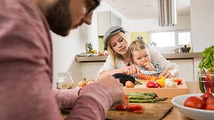 Mutter, Vater und Kind kochen gemeinsam in der Küche. Sie sitzen am Küchentisch und bereiten das Gemüse vor.