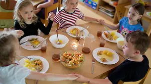 Kinder sitzen an einem kleinen Holztisch zum Mittagessen in einer Kita.