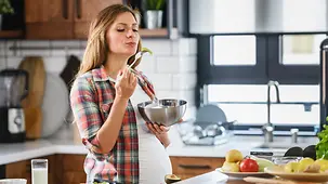 Schwangere Frau steht in der Küche und kostet ihr Essen.
