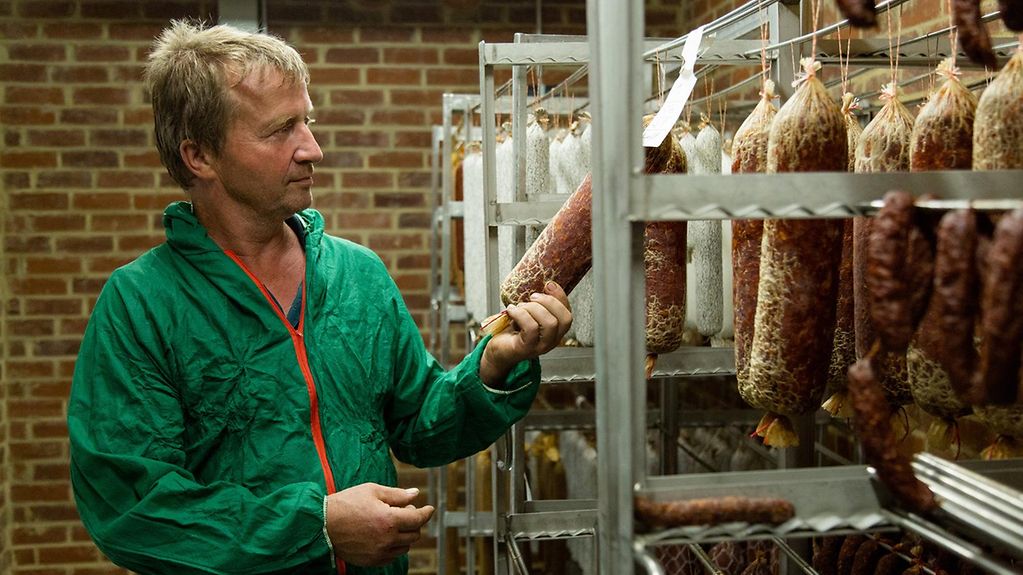 Landwirt Christopf Leiders steht mit einer Hygieneschutzkleidung in seiner Metzgerei und begutachtet eine Salami.