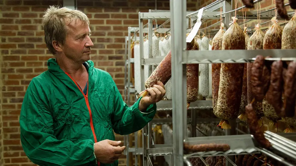 Landwirt Christopf Leiders steht mit einer Hygieneschutzkleidung in seiner Metzgerei und begutachtet eine Salami.