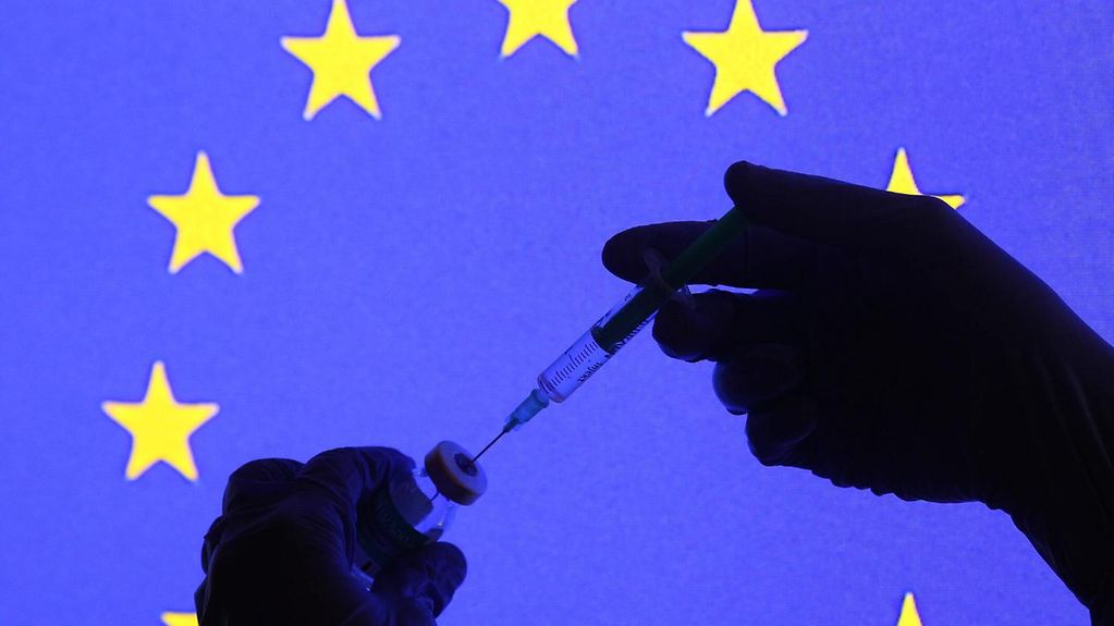 EU flag, in front the silhouette of a coronavirus syringe and a hand wearing a rubber glove