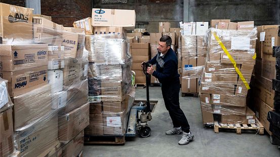 Ein Mitarbeiter transportiert in einem Warenlager eine mit Kartons beladene Palette.
