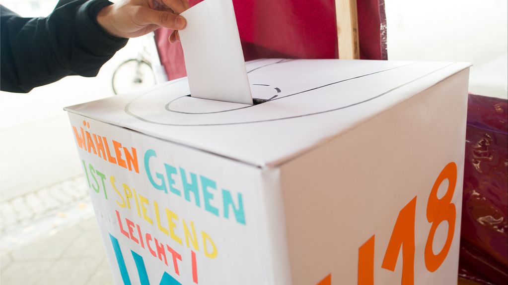 In eine bunt bemalte Wahlurne wird ein Wahlzettel eingeworfen. Auf der Urne steht "Wählen gehen ist spielend leicht!"