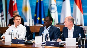 Der französische Präsident Emmanuel Macron, Macky Sall Präsident des Senegal und Vorsitzender der Afrikanischen Union (AU) und Bundeskanzler Olaf Scholz (v.l.) am Konferenztisch zu Beginn der fünften Arbeitssitzung.