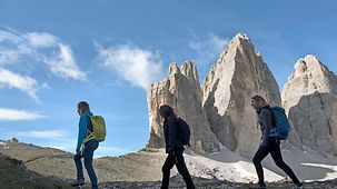 Drei Bergwanderinnen gehen auf einem Grat vor den Drei Zinnen vorbei.