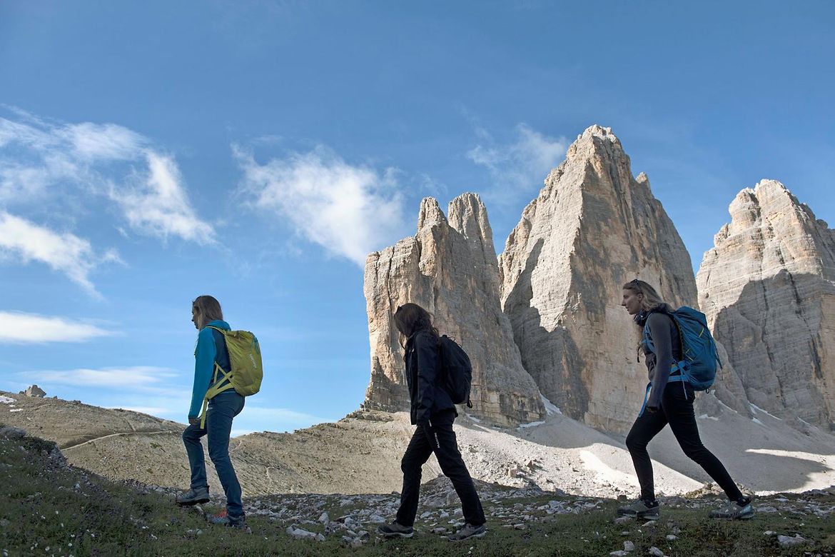 Drei Bergwanderinnen gehen auf einem Grat vor den Drei Zinnen vorbei.