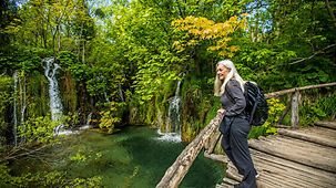 Eine ältere Wanderin blickt von einem hölzernen Steeg auf einen See, in den sich kleine Wasserfälle ergießen.