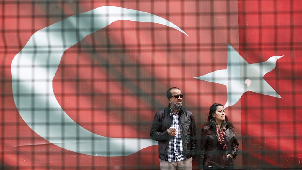 Two people stand in front of an enormous Turkish flag.