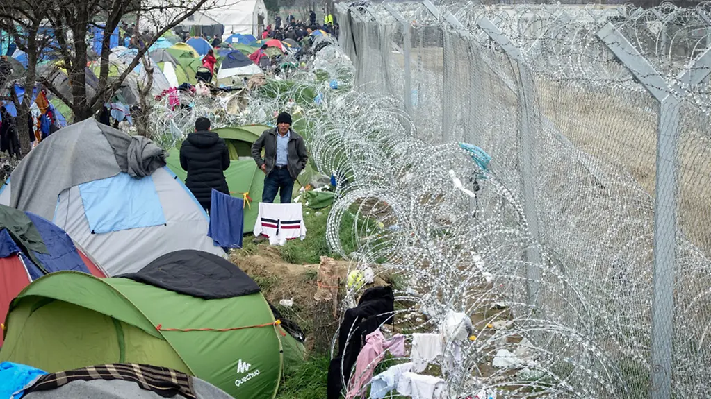 Flüchtlinge stehen am im Flüchtlingslager in Idomeni an der Grenze zwischen Griechenland und Mazedonien vor ihren Zelten am mit Stacheldraht gesicherten Grenzzaun.