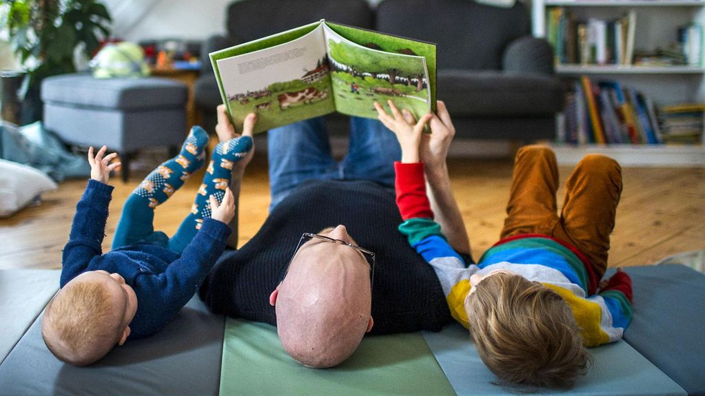 Ein Vater liest mit seinen beiden Kindern auf dem Boden liegend ein Buch