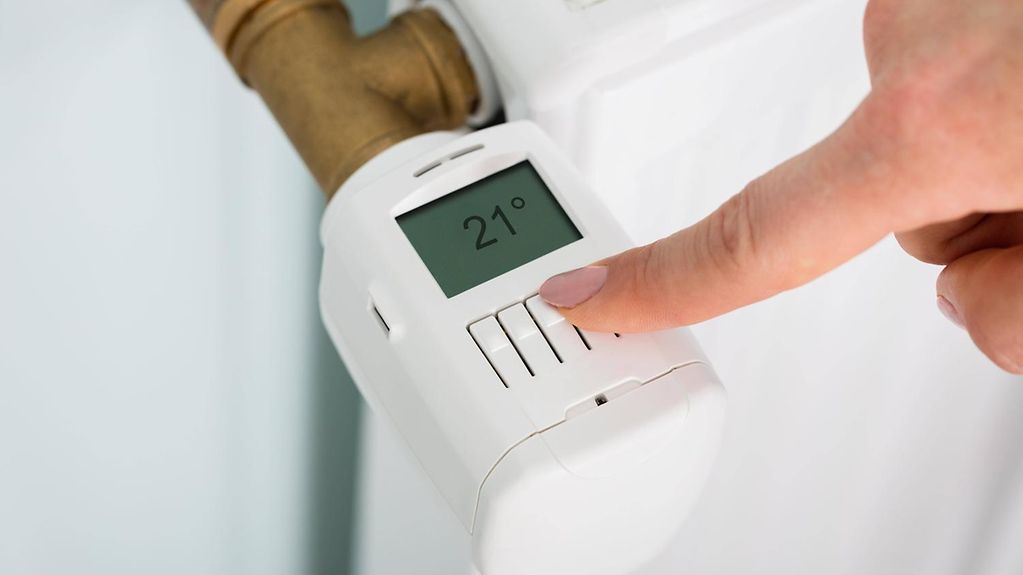 Eine Person stellt ein digitales Thermostat an einer Heizung auf 21 Grad Celsius.