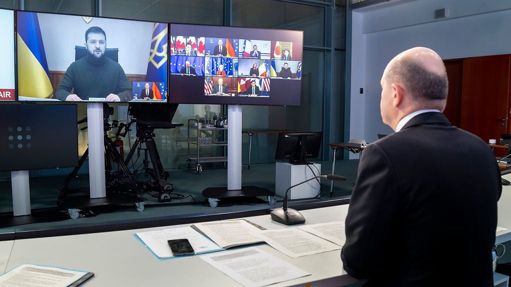 Auf dem Foto ist Kanzler Scholz vor einem Bildschirm zu sehen, auf dem gerade der ukrainische Präsident Selenskyj spricht.