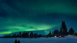  Zwei Menschen sitzen auf dem Eis eines Sees und beobachten einen Nachthimmel mit Polarlichtern in Lappland im Norden Schwedens.