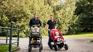 Zwei Väter mir ihren Kindern im Kinderwagen beim Spazieren im Park.