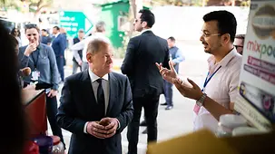 Bundeskanzler Scholz hält enen Teebecher in der Hand während der Teestandbesitzer mit ihm spricht. 
