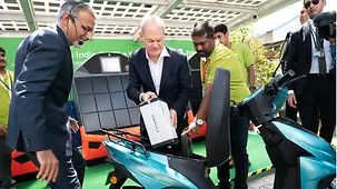 Bundeskanzler Scholz steht mit zwei Mitarbeitern des Unternehmens vor einem E-Roller und hält die Antriebsbatterie in den Händen. 