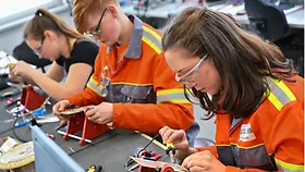 Drei junge Auszubildene in gelber Arbeitskleidung an einer Elektrowerkbank. 