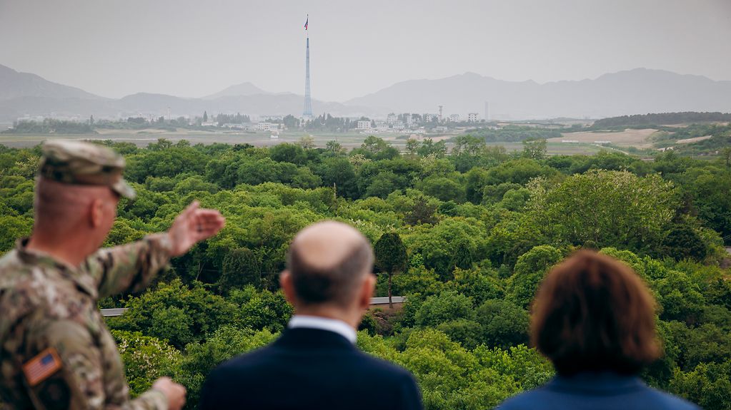 Ein Soldat zeigt in Richtung einer Landschaft. Hinter Bäumen sind Gebäude und ein riesiger Flaggenturm mit der nordkoreanischen Flagge zu sehen. Von hinten sind Bundeskanzler Olaf Scholz und seine Frau Britta Ernst zu erkennen.