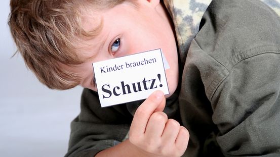 Kind hält Zettel mit der Aufschrift "Kinder brauchen Schutz" in der Hand.