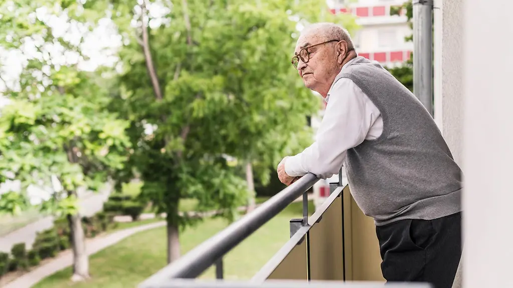 EIn älterer Mann steht alleine auf dem Balkon und schaut in die Umgebung.