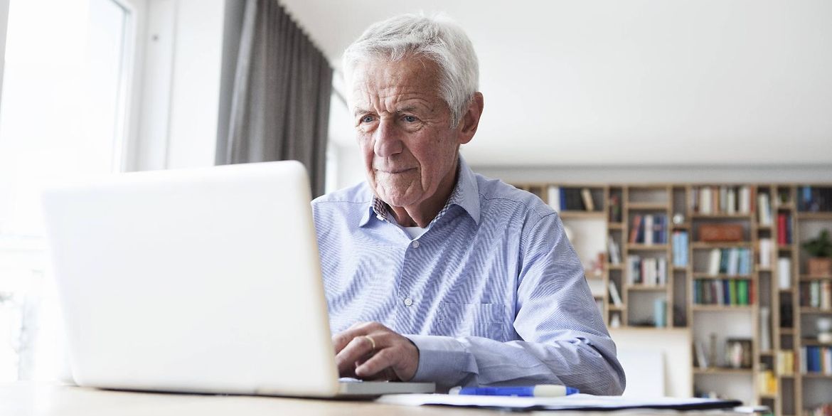 Porträt eines älteren Mannes, der am Tisch sitzt und einen Laptop benutzt. 