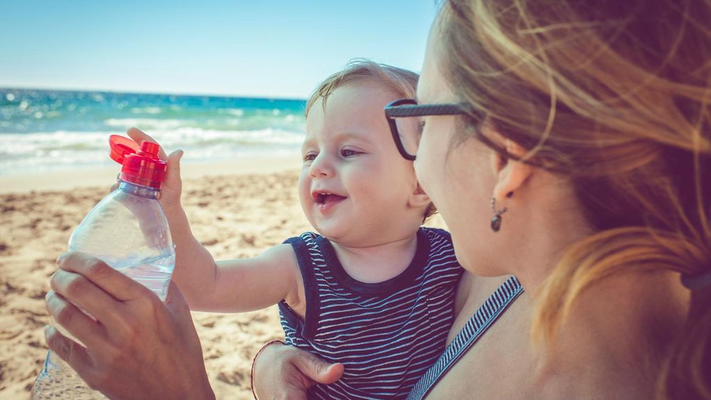Eine Frau hält ein Kleinkind auf dem Arm. Sie sind am Strand. Die Frau hält eine Wasserflasche in der Hand. 
